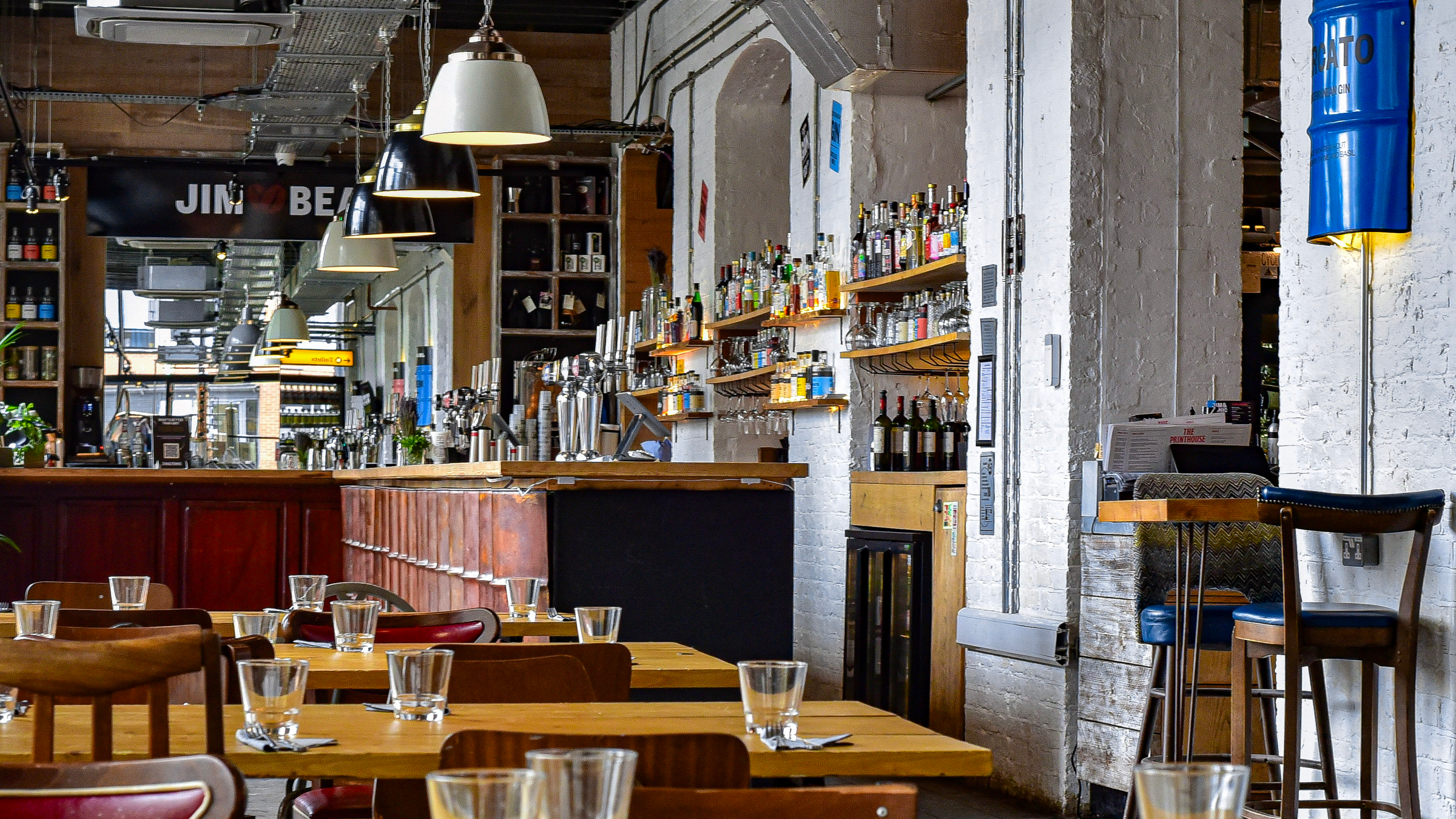 A photo of a classy bar that is full of all sorts of alcohol with chairs and tables with glasses on in the foreground.