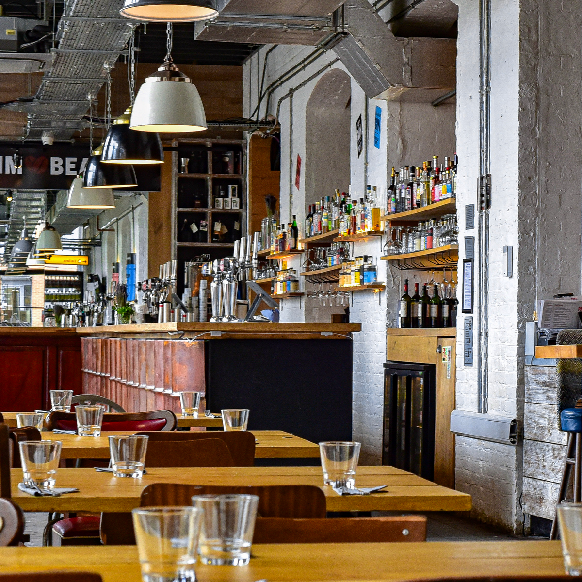 A photo of a classy bar that is full of all sorts of alcohol with chairs and tables with glasses on in the foreground.