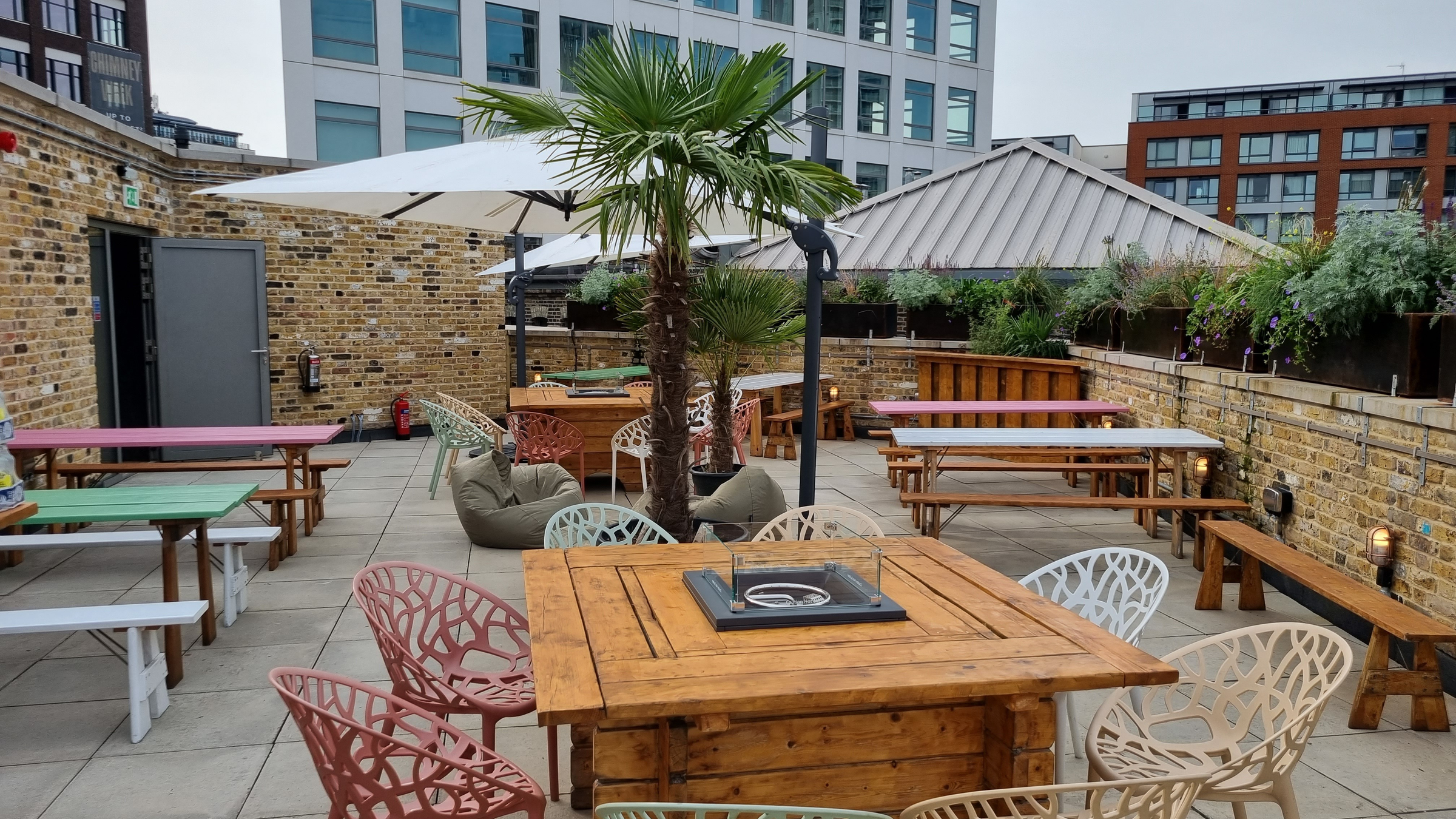 An outdoor seating area on a large balcony with picnic benches and tables surrounded by varyingly coloured chairs.