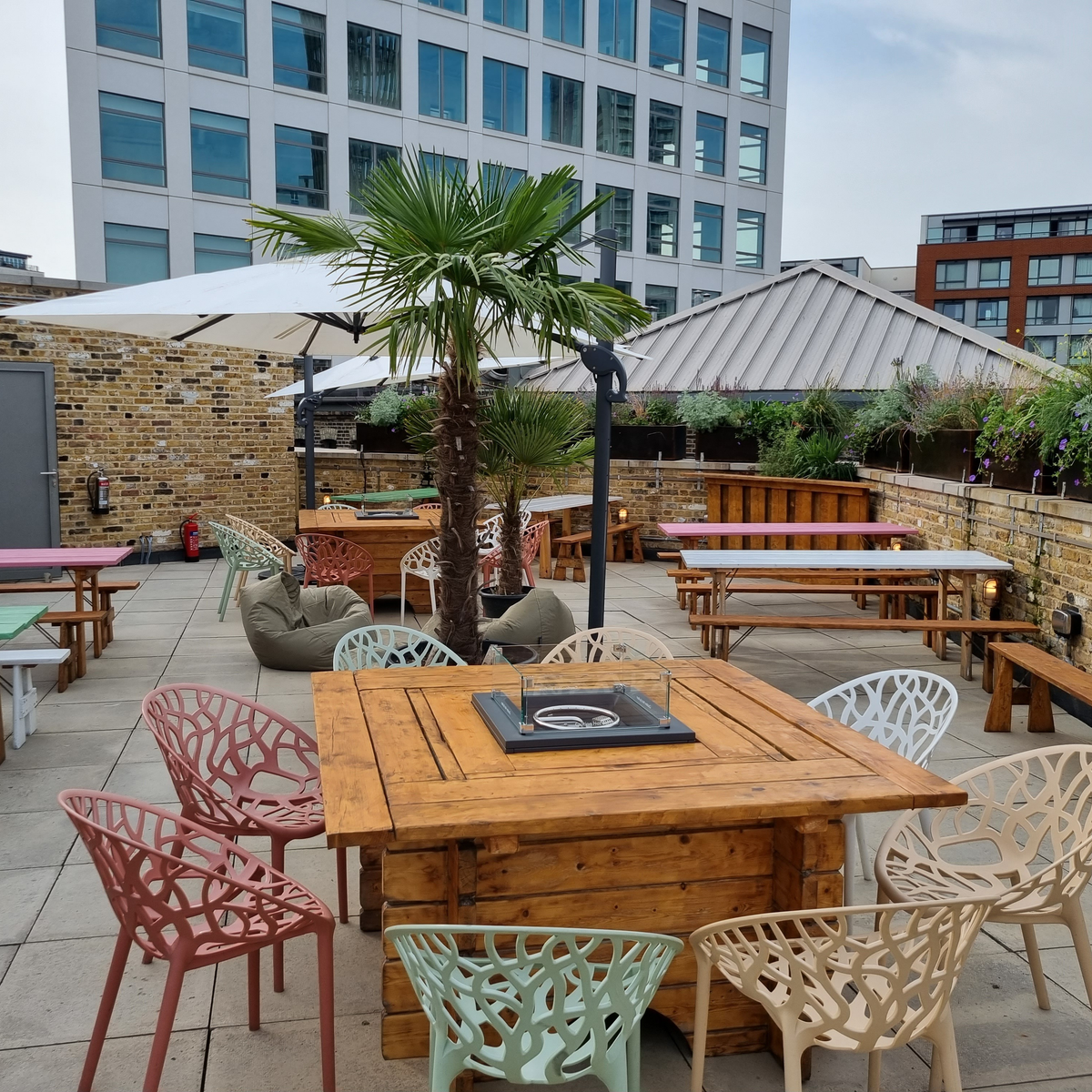 An outdoor seating area on a large balcony sporting picnic benches and tables surrounded by chairs of varying colours.