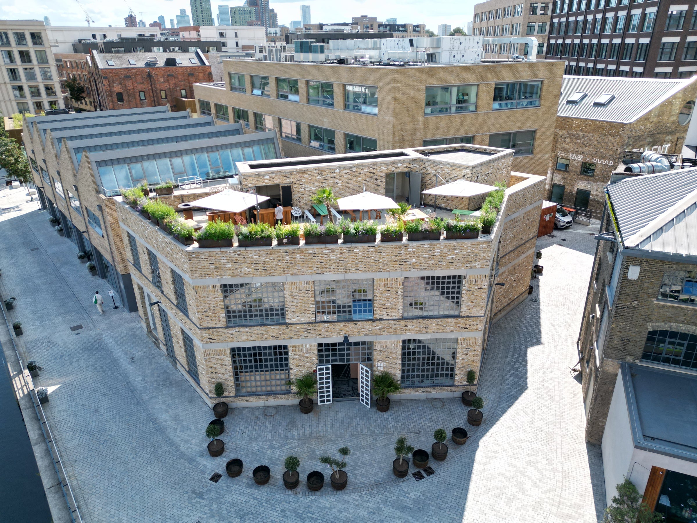 An overhead view of a brick building with large windows and a rooftop garden terrace, set within an industrial-style courtyard surrounded by other commercial buildings. The terrace features seating areas, greenery, and umbrellas.
