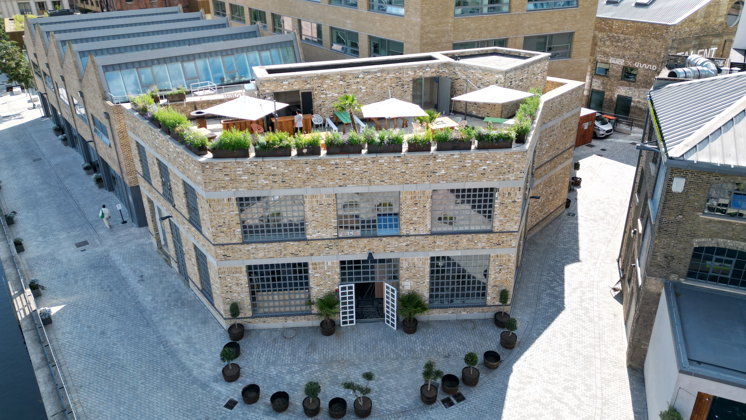 An overhead view of a brick building with large windows and a rooftop garden terrace, set within an industrial-style courtyard surrounded by other commercial buildings. The terrace features seating areas, greenery, and umbrellas.