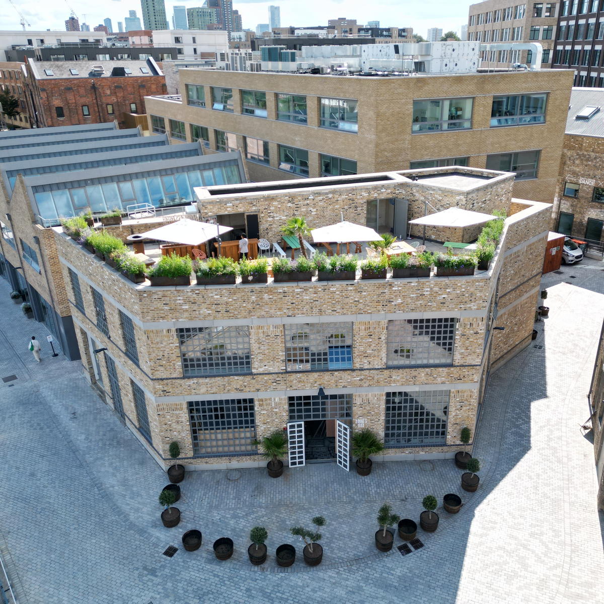 An overhead view of a brick building with large windows and a rooftop garden terrace, set within an industrial-style courtyard surrounded by other commercial buildings. The terrace features seating areas, greenery, and umbrellas.
