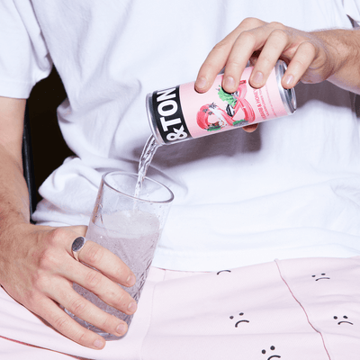 A man in a plain white tee pouring a can of Jim and Tonic Rhubarb and Honey gin from a pink can into a clear glass.