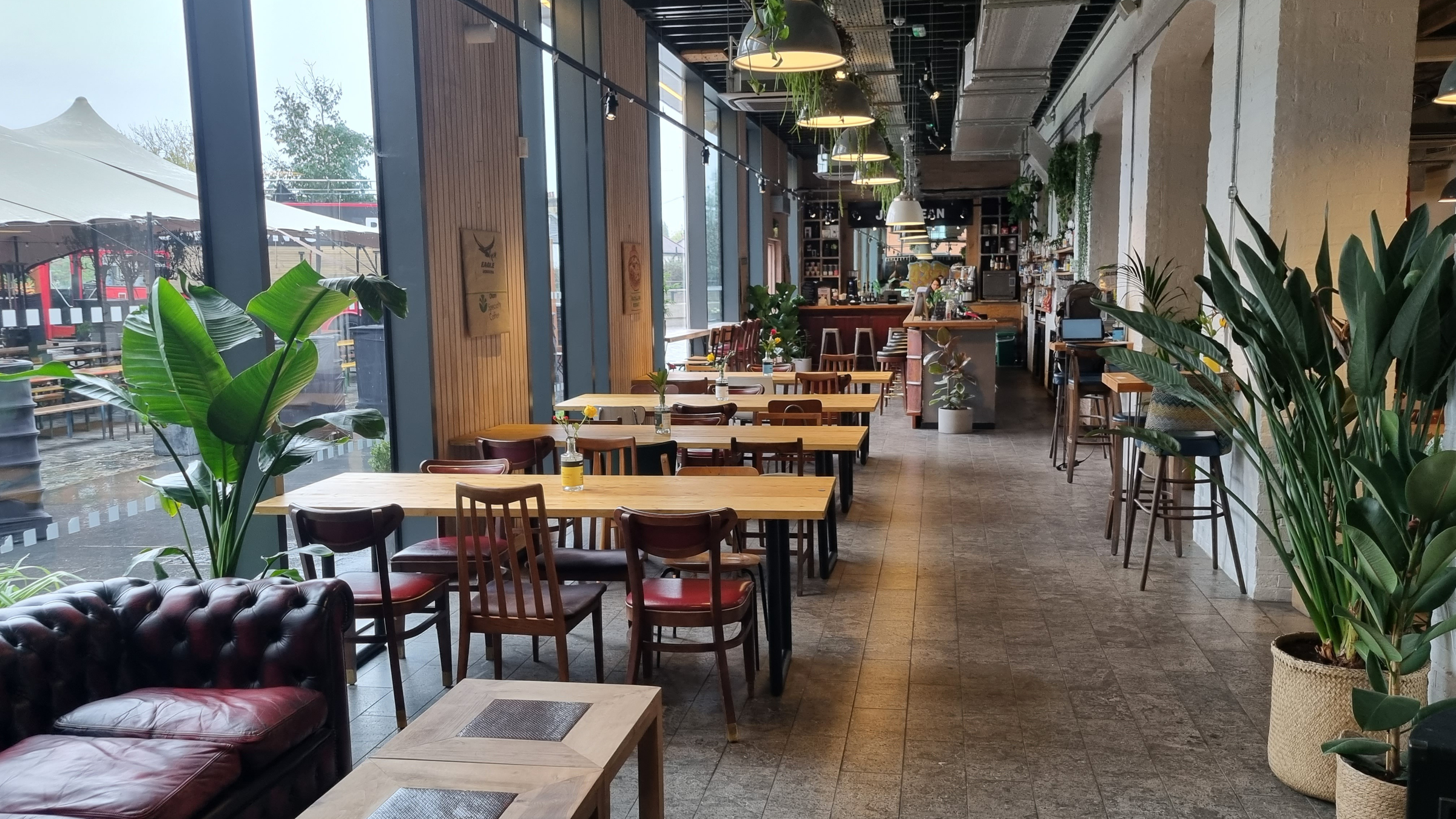 An inviting interior with wooden tables, a leather sofa, and large windows. The space is filled with greenery and has a cozy atmosphere.