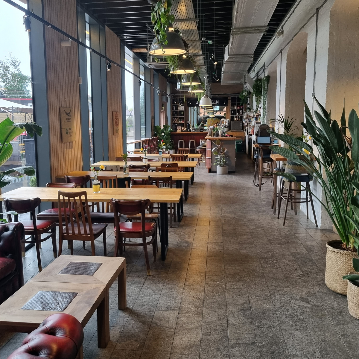 An inviting interior with wooden tables, a leather sofa, and large windows. The space is filled with greenery and has a cozy atmosphere.