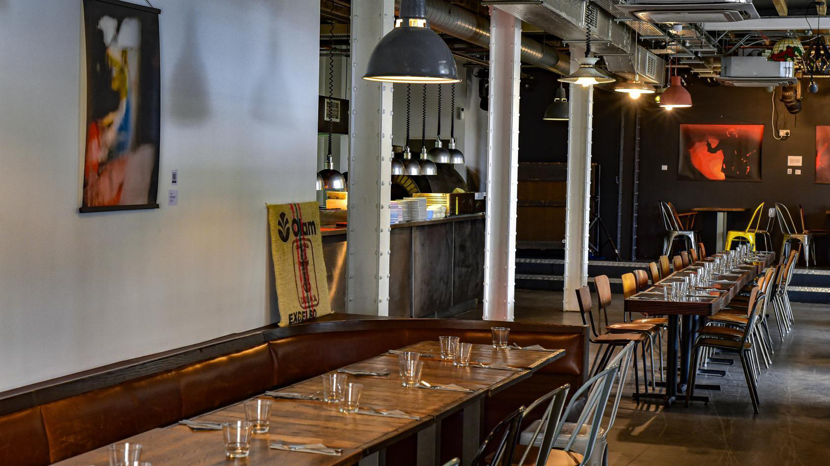 An inviting bar restaurant showcasing a long table adorned with clear glasses and plates. The industrial décor features metal accents, and the walls are decorated with vibrant artwork.